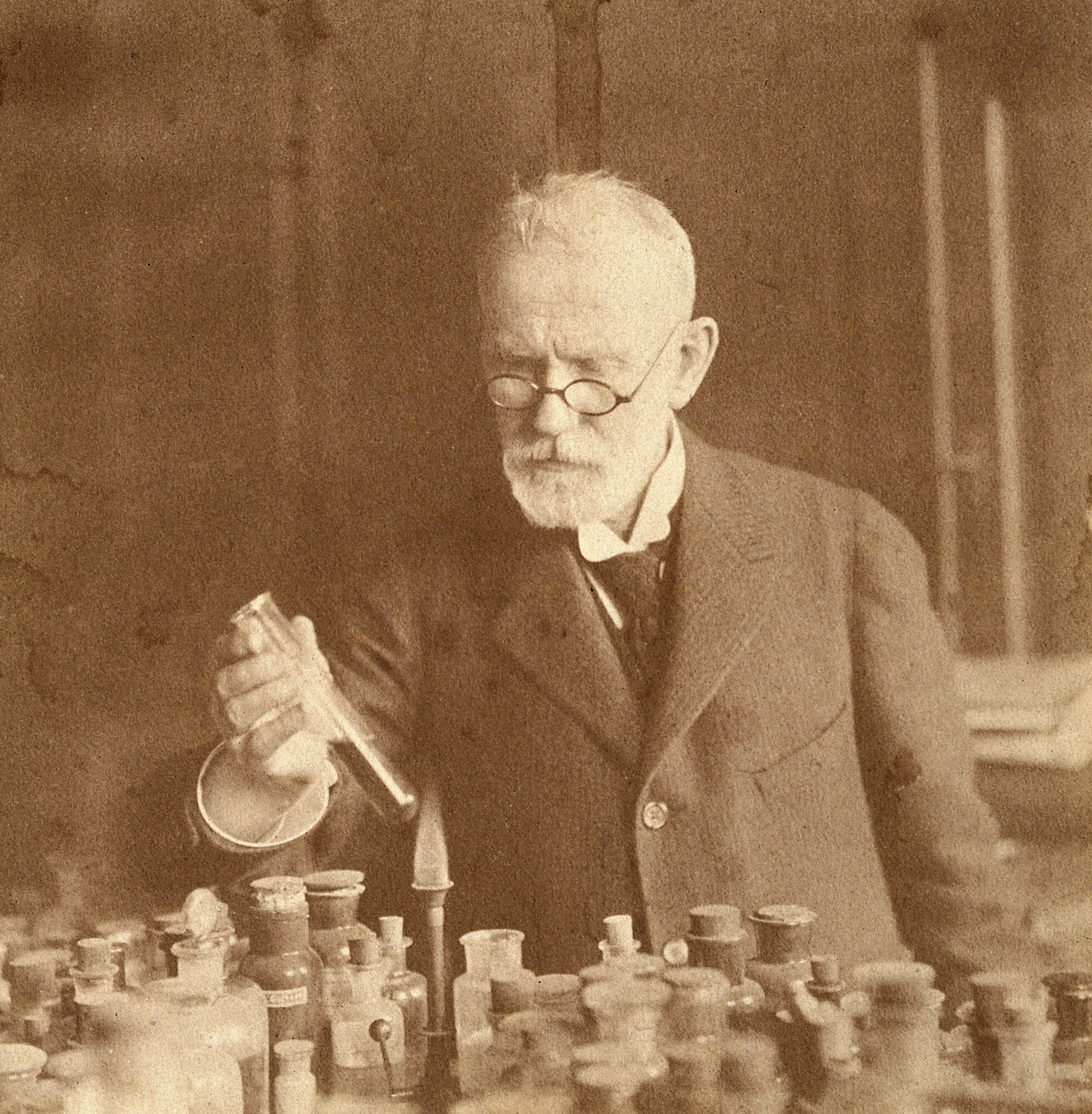 Sepia-toned photograph of Paul Ehrlich in a laboratory, holding a glass tube. He is standing behind a table filled with glass bottles and scientific apparatus.