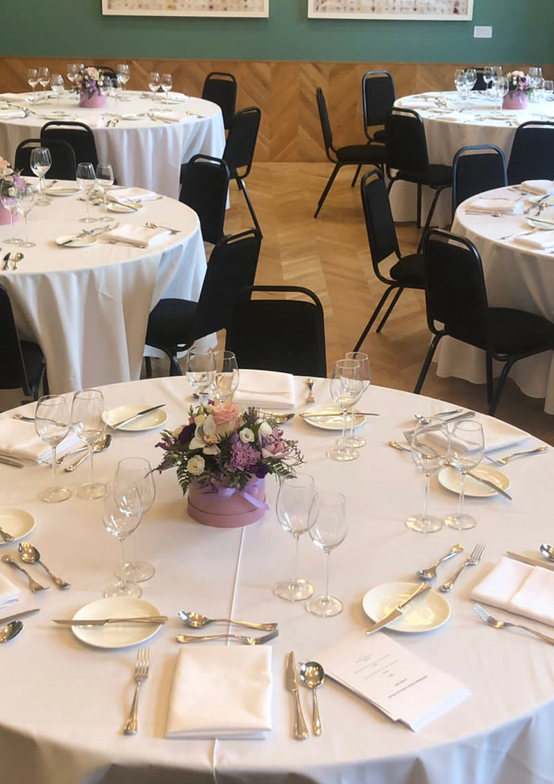Event room set up banquet-style with round dining tables, white tablecloths, floral centrepieces and place settings for a formal meal.