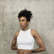 Black and white photo of a young person leaning against a wall with their hands behind their back. They are looking straight at the camera.
