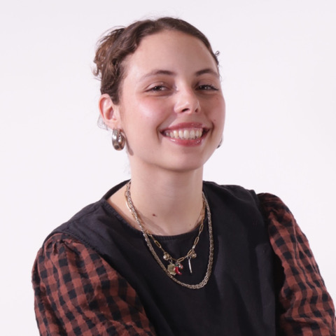 Headshot of a person with tied-back brown hair, wearing hoop earrings, layered necklaces and a black top with checked sleeves, against a plain background.
