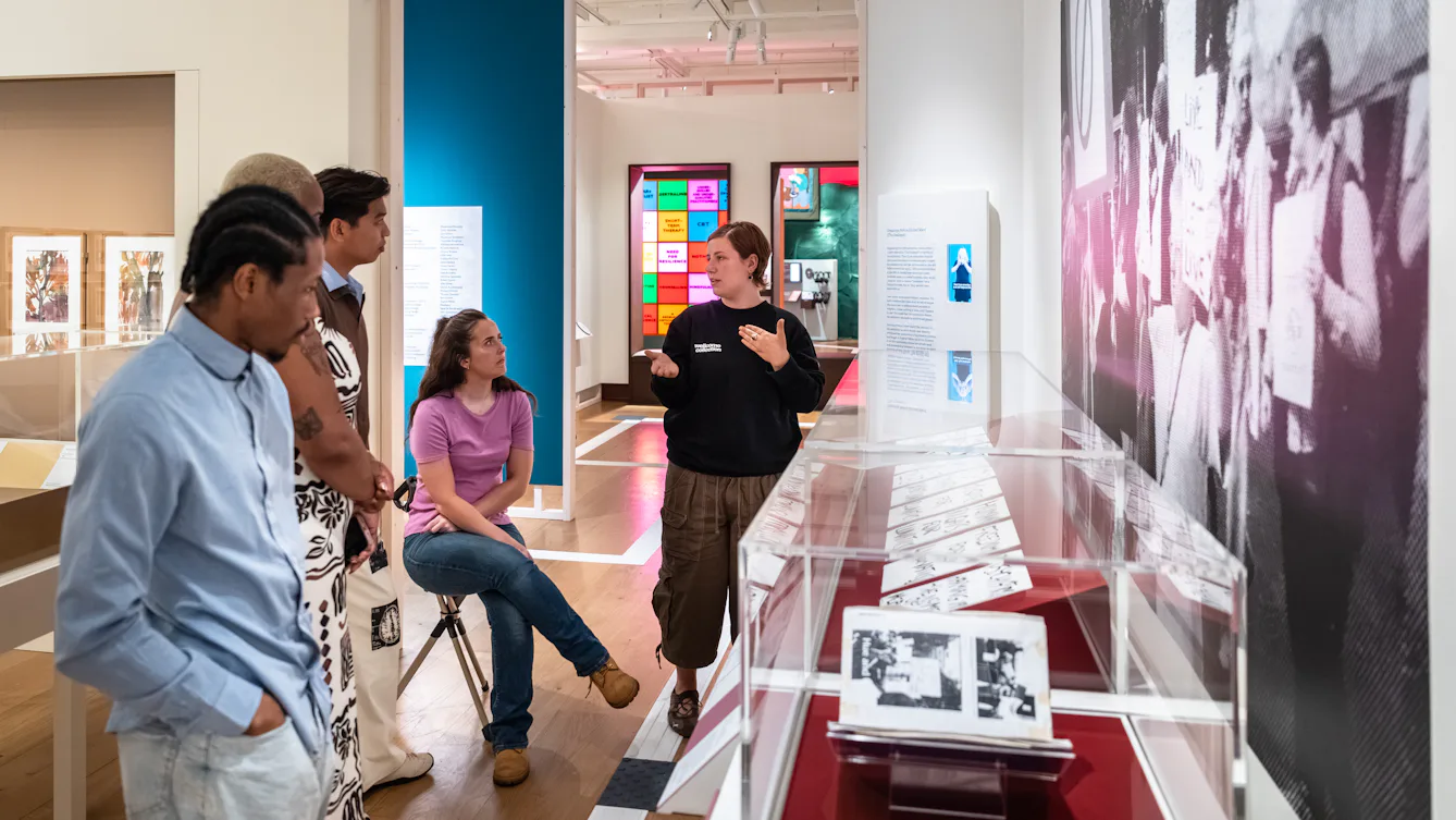 Photograph of a Visitor Experience and Engagement Facilitator giving a guided tour of a colourful gallery space to a group of 4 visitors.