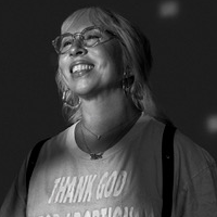 Black-and-white headshot of a person with short hair and glasses, wearing a T-shirt and necklace against a dark background.