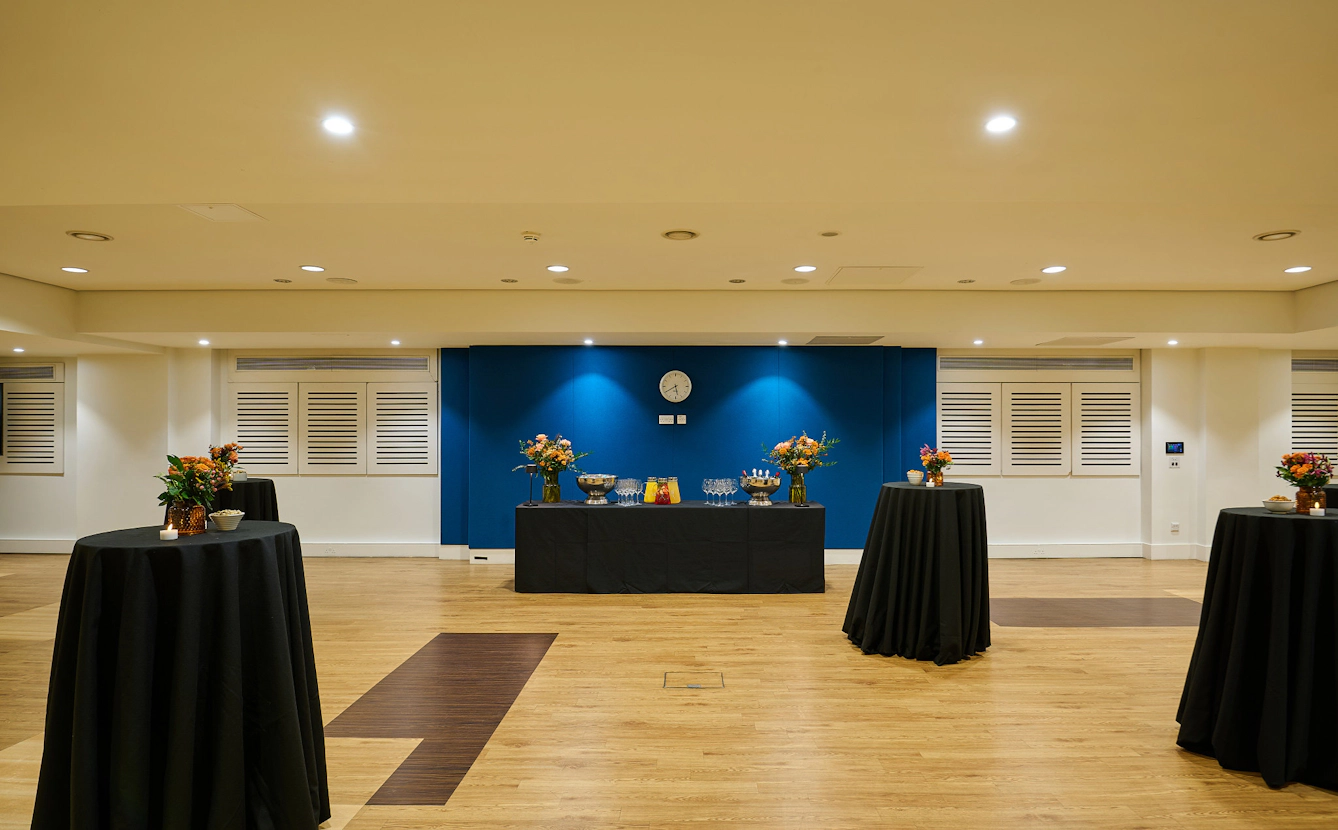 Open event space with wooden flooring, cocktail tables and a central refreshments station, set up for a drinks reception or networking event.