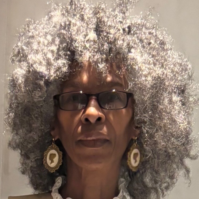 Headshot of a person with curly grey hair, dark-framed glasses and large round earrings, against a plain background.