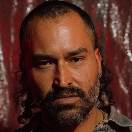 Close-up headshot of a person with short hair, a full beard, nose ring and earring, against a textured red background.