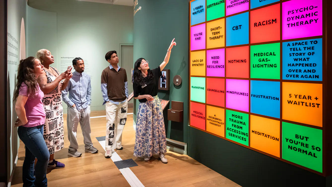 Photograph of a Visitor Experience and Engagement Facilitator giving a guided tour of a colourful gallery space to a group of 4 visitors.
