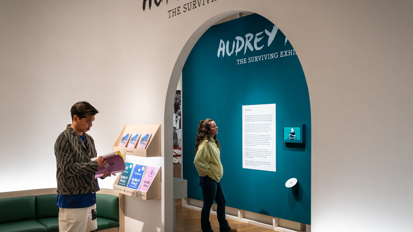A view of the entrance to 'Audrey Amiss: The Surviving Exhibitions'. A visitor picks up a gallery guide while another visitor reads exhibition text through an archway.