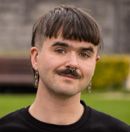 Black and white photo of the head and shoulders of a young person with short dark hair and a moustache. They are smiling and looking directly at the camera.
