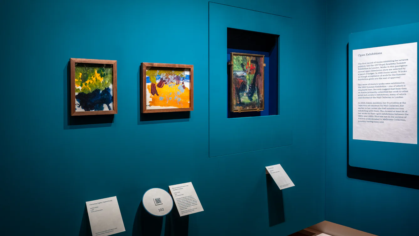 A view of a room in the exhibition with three framed paintings by Audrey Amiss displayed on a blue gallery wall, with QR codes and labels underneath. On the right is the exhibition wall text.