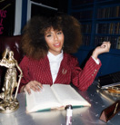 A smartly dressed woman sits at a desk with a large open book infront of her. She is smiling and looking at the camera.