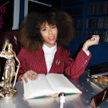 A smartly dressed woman sits at a desk with a large open book infront of her. She is smiling and looking at the camera.