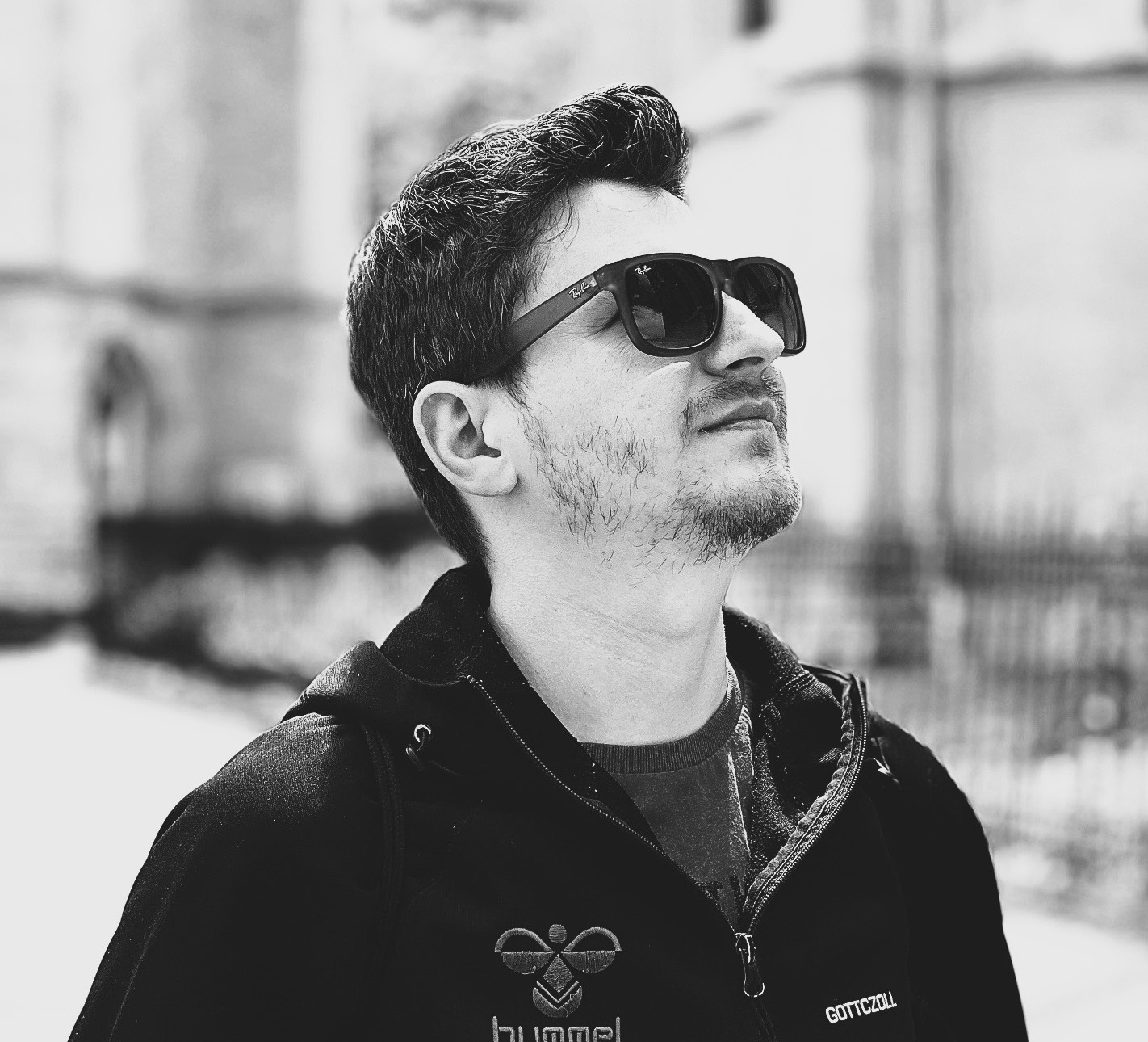 Black and white photograph of the head and shoulders of a young man wearing dark sunglasses and looking up to the top right of the image.