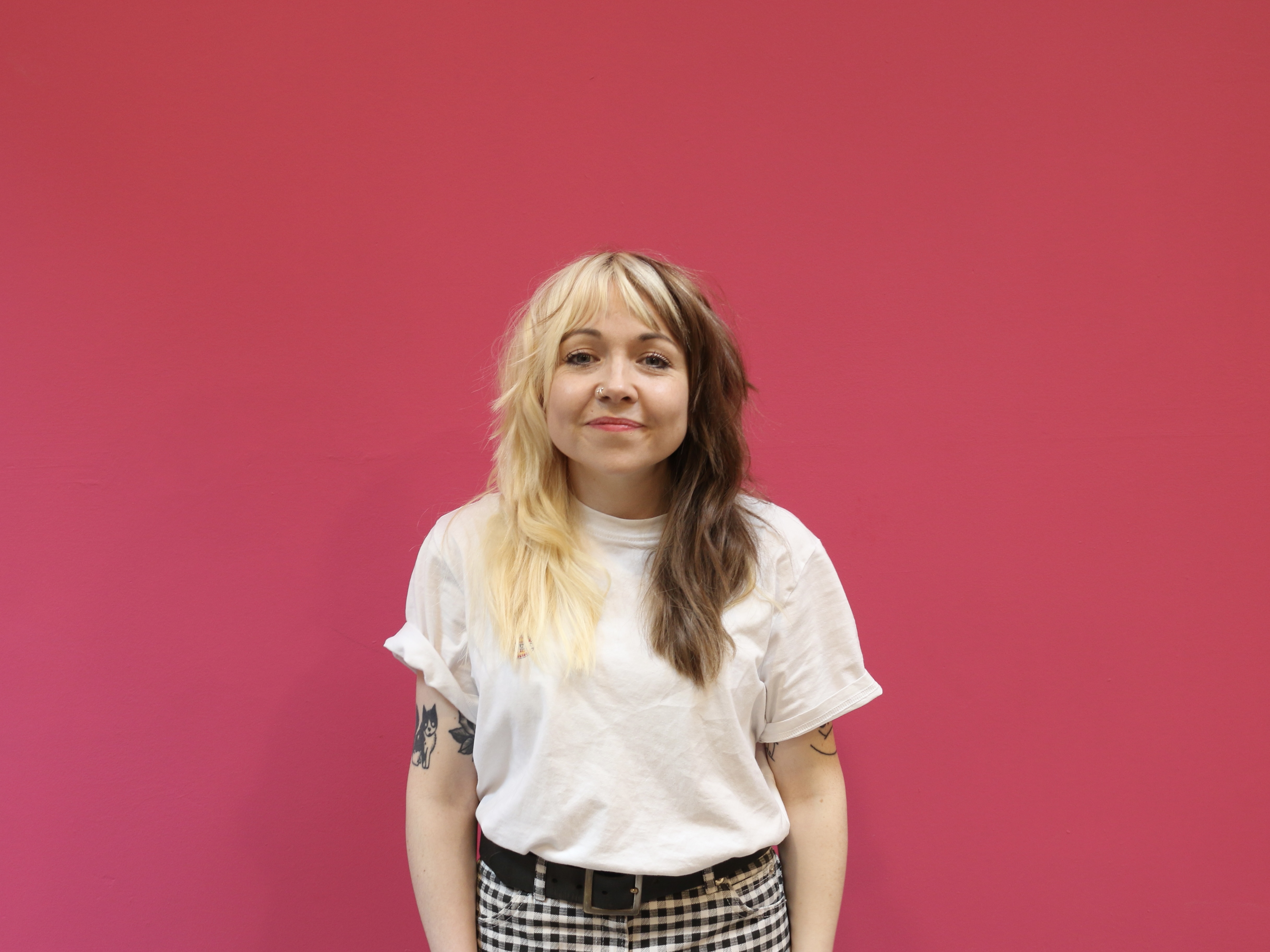 Head and shoulder shot of visual artist Rose Sergent who is smiling to camera, has long hair that is half blonde, half brunette and is wearing a white top and checked trousers.