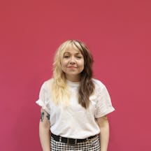 Head and shoulder shot of visual artist Rose Sergent who is smiling to camera, has long hair that is half blonde, half brunette and is wearing a white top and checked trousers.