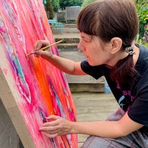 An artist with shoulder length, brown hair tied into pigtails who is painting on a large canvas.