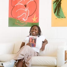 Zine-maker Zoë is sat cross-legged on a sofa, holding a zine and smiling to camera.