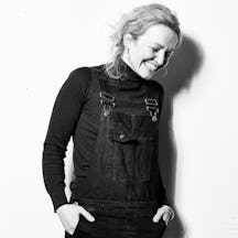 Photo of artist Lucy wearing dungarees with their hands in their pockets, shoulder length wavy hair and a big smile across their face.