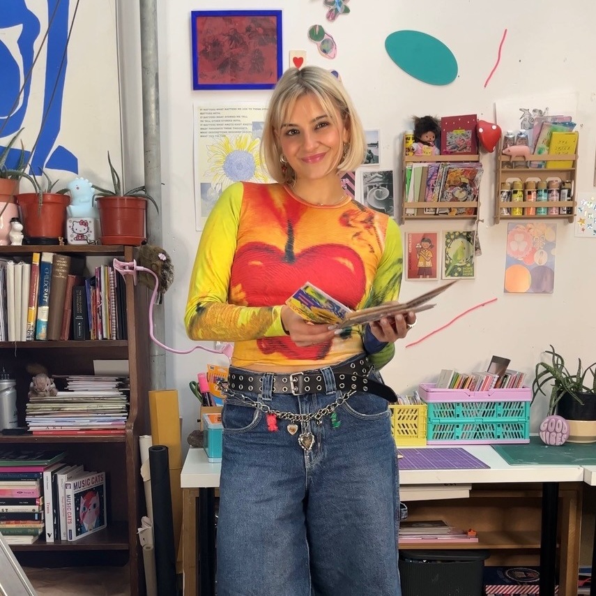 Photo of artist Ioana smiling to camera and standing in an art studio, holding a zine, surrounded by paints and crafting materials.