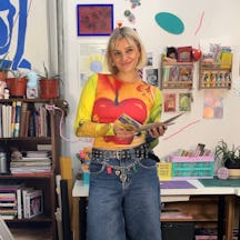 Photo of artist Ioana smiling to camera and standing in an art studio, holding a zine, surrounded by paints and crafting materials.