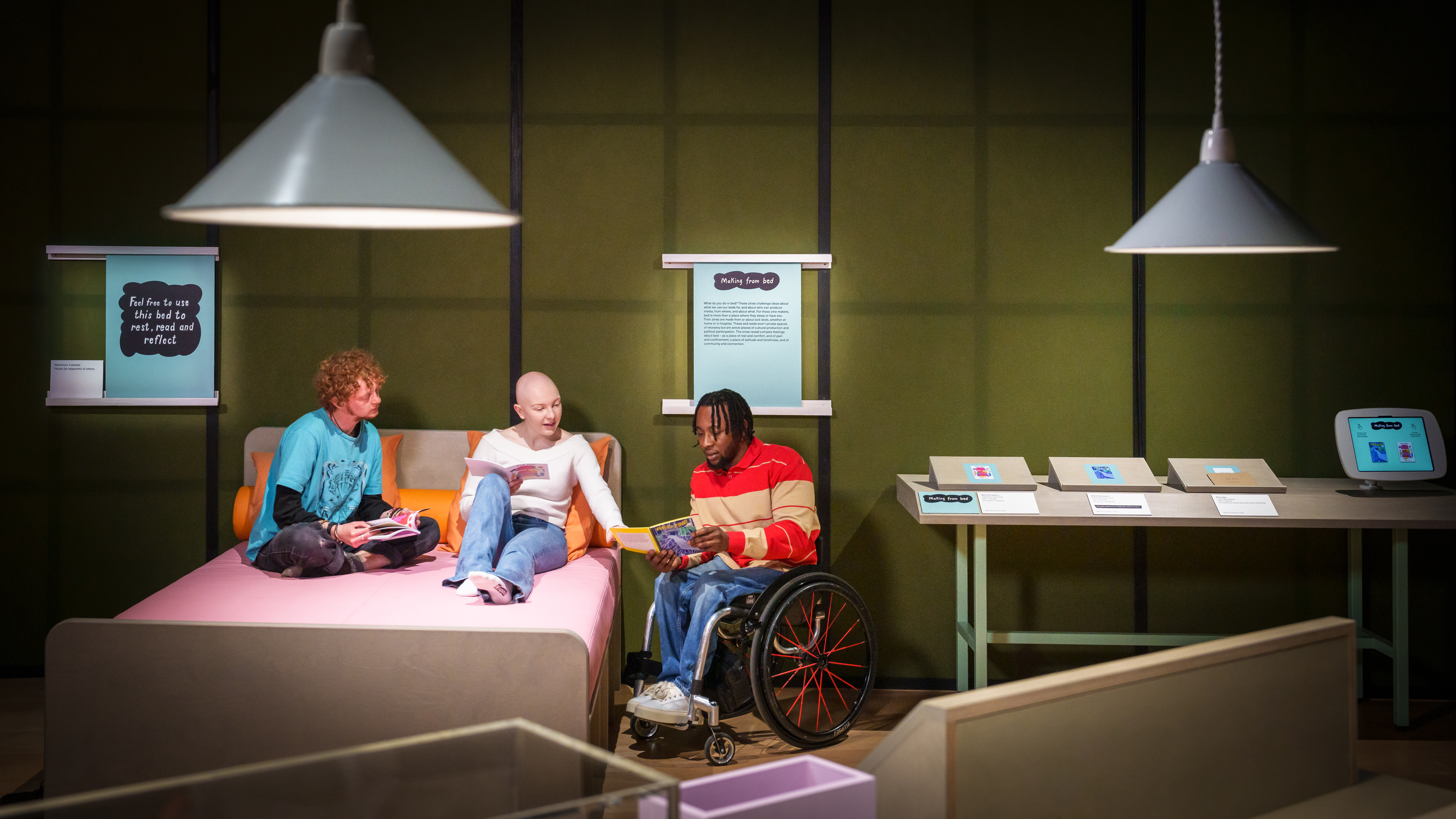 Photograph of three people chatting in an exhibition gallery. In the centre one of the people is discussing a zine they have held open in their hands with the other two people, who are sat on a large double bed, made up of a pink mattress and orange pillows. To the side of the bed is a display table with more zines and a tablet in a holder.