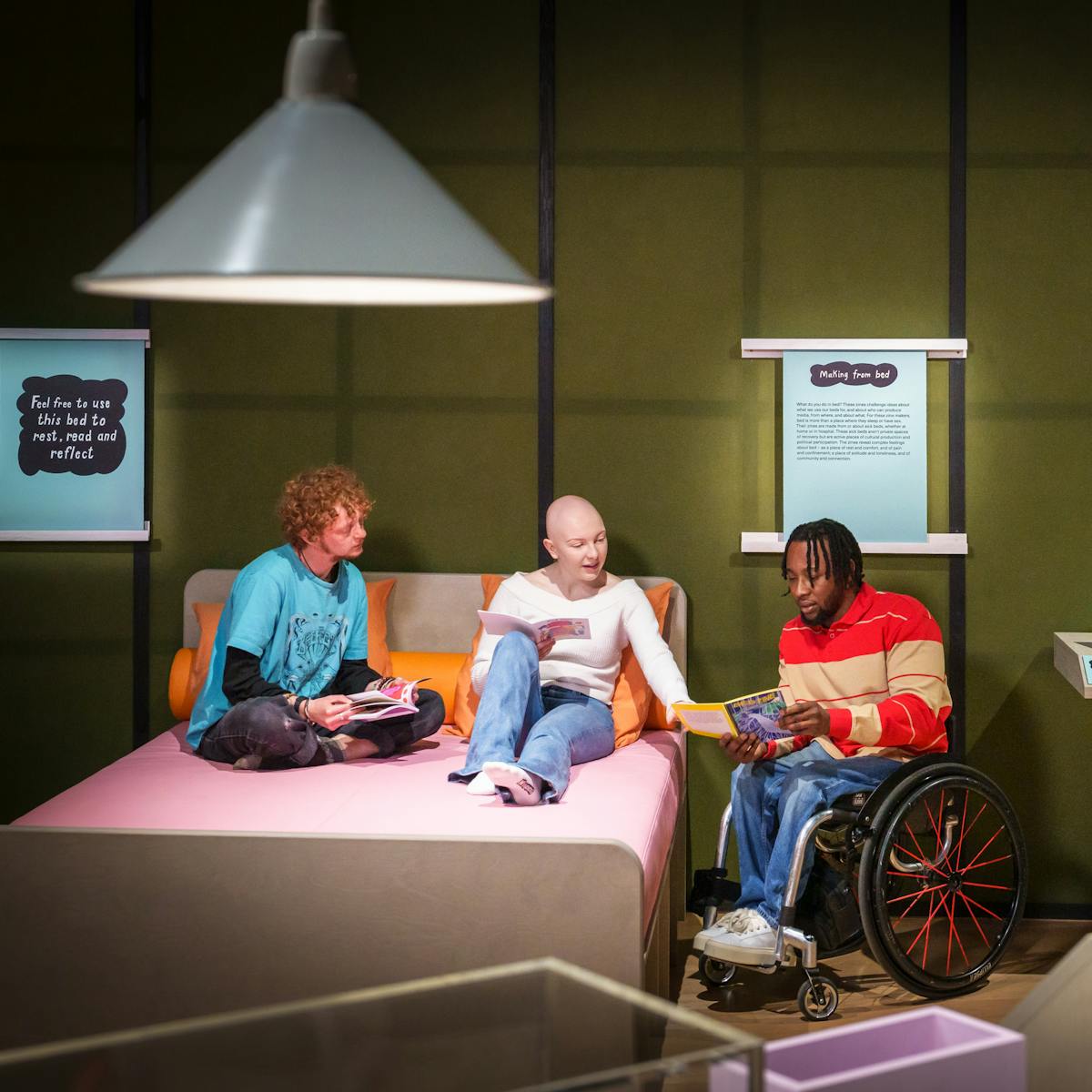 Photograph of three people chatting in an exhibition gallery. In the centre one of the people is discussing a zine they have held open in their hands with the other two people, who are sat on a large double bed, made up of a pink mattress and orange pillows. To the side of the bed is a display table with more zines and a tablet in a holder.