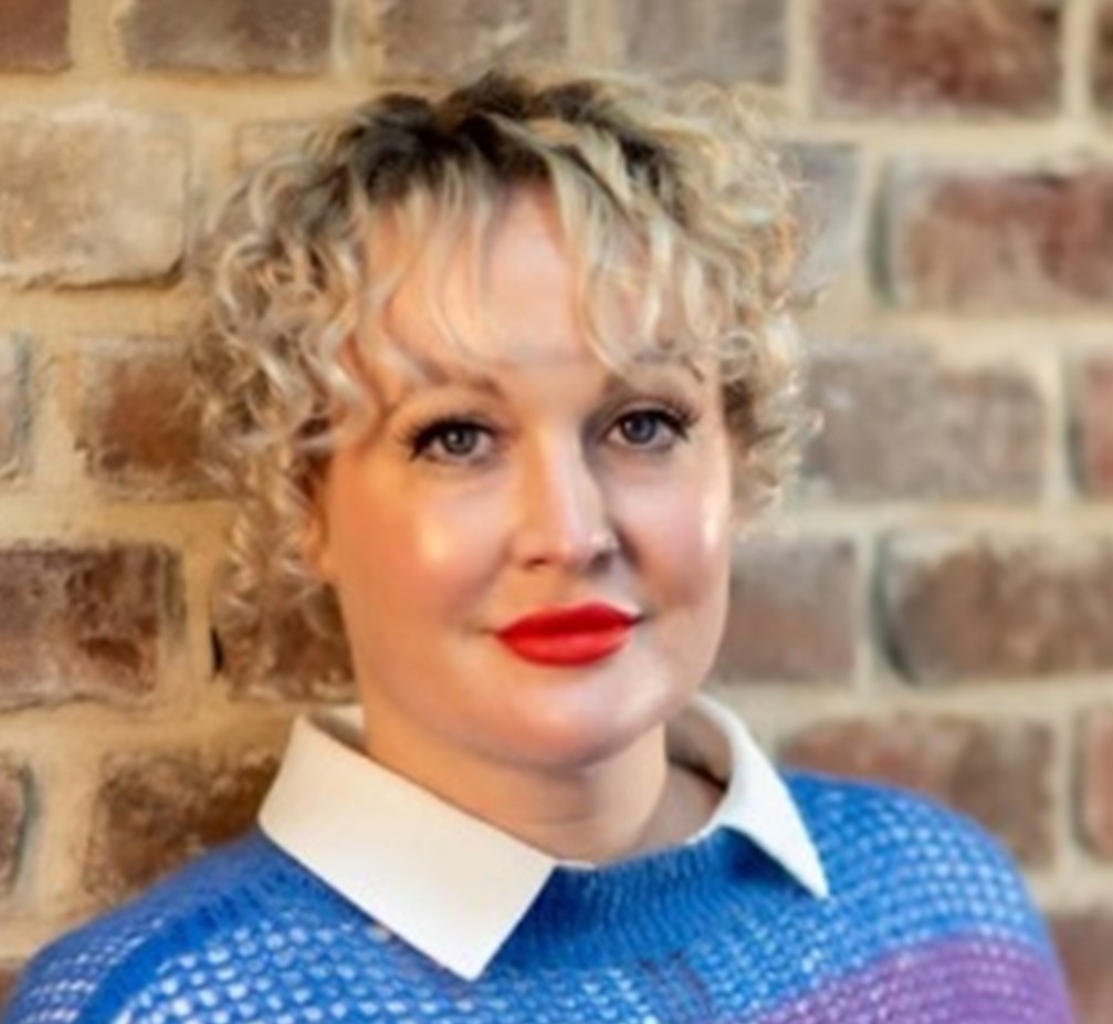 A headshot of Kate Lister, who has short blonde curly hair, is wearing a blue jumper and is leaning against a brick wall.