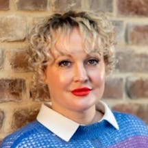 A headshot of Kate Lister, who has short blonde curly hair, is wearing a blue jumper and is leaning against a brick wall.