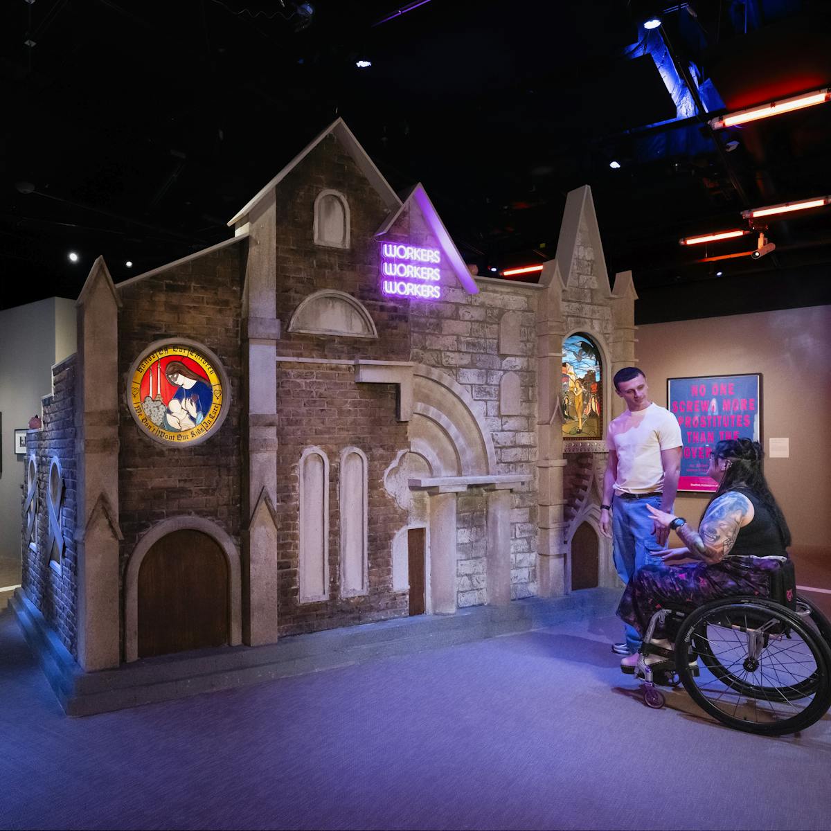 A photograph of two people in an exhibition space looking at a large scale installation artwork. The artwork consists of the facade of a church with stained glass windows depicting sex workers and a pink neon sign of the of the words workers, workers, workers. In the background another person is watching a film screening and wearing headphones.