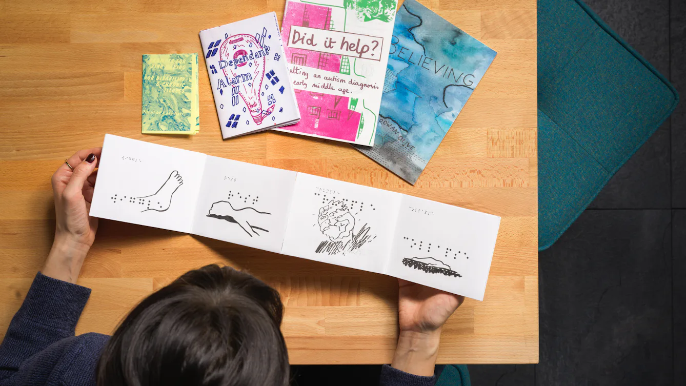 A selection of 5 zines arranged on a table as seen from above the table. A person sits at the table with one of the zines open infront of them.