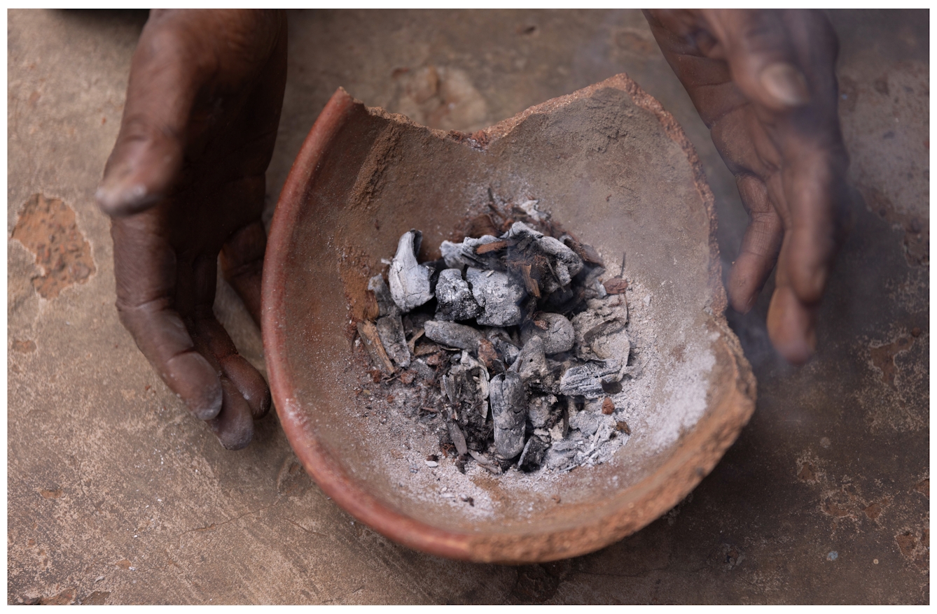 A pair of hands holding a broken clay bowl filled with burnt kola nuts, ashes and small charcoal pieces, resting on a rough, brown surface.