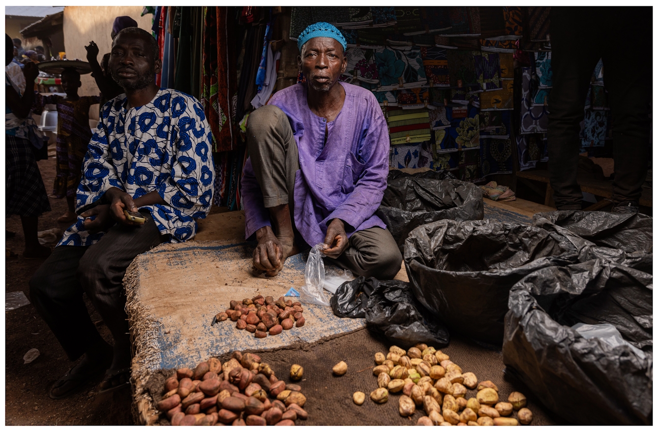 A man sits in his market stall wearing a blue hat and purple shirt. In front of him are arrangements of kola nuts which he is selling. Next to him is another man wearing a patterned blue shirt, and in the background are bright fabrics and other market activity.