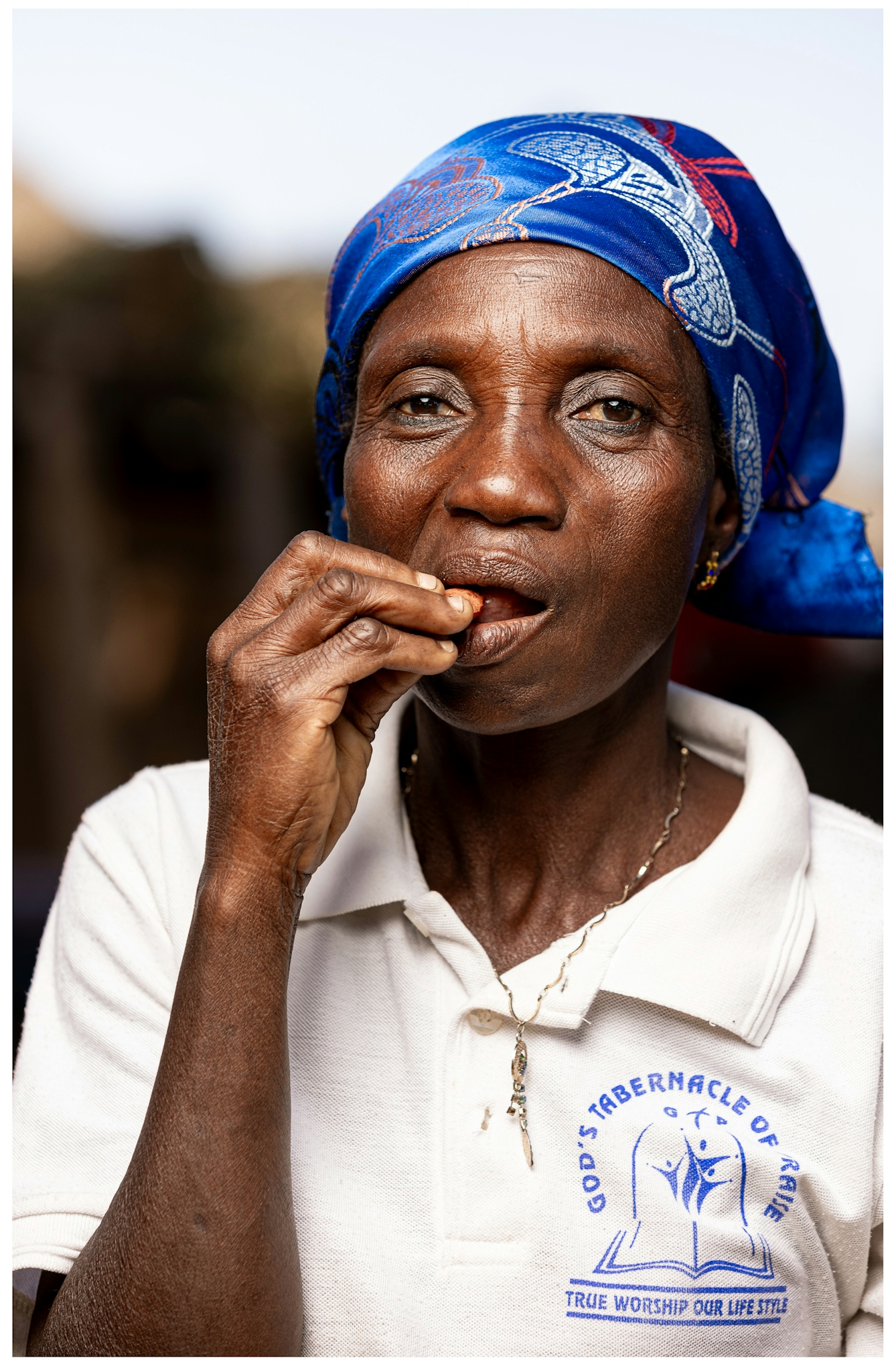 A woman wearing a blue headscarf and a white shirt is holding her hand to her mouth and taking a bite of a kola nut. The background is softly blurred.