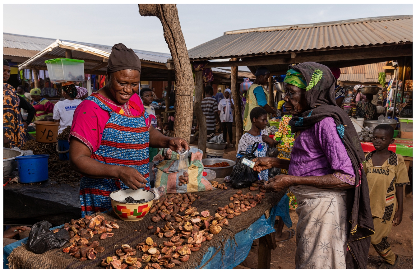 Two women at an outdoor market stall engage in a transaction. One woman bags kola nut while the other, holds money in her hand. Various products and people fill the bustling market, under a corrugated metal roof.