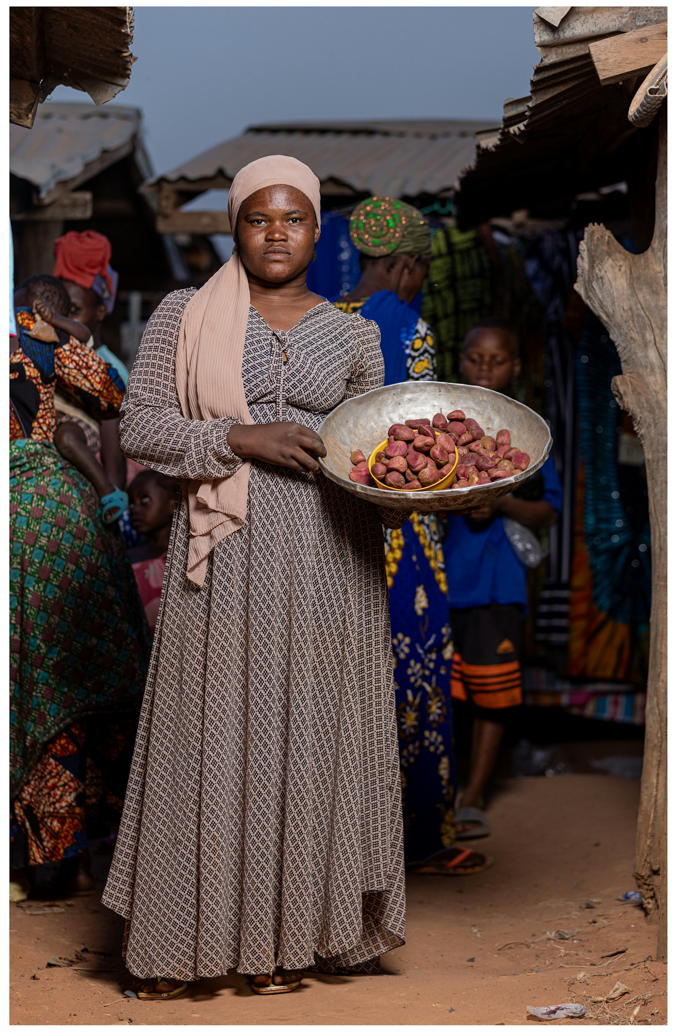 A woman wearing a patterned dress and headscarf stands in a market, holding a large bowl of reddish-brown kola nuts. She is surrounded by people and stalls with colorful fabrics in the background.