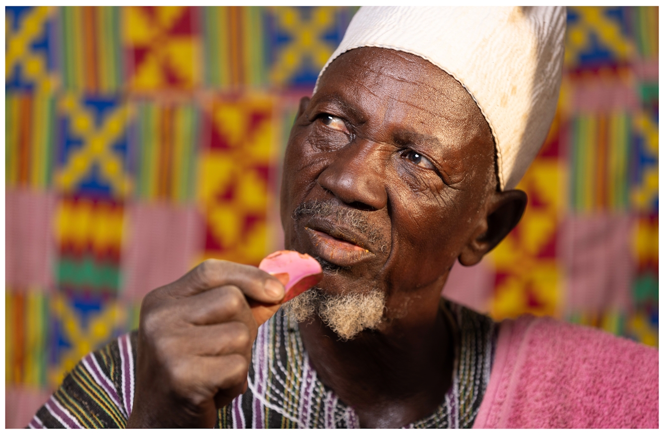 A man wearing a traditional hat and striped clothing eats a pink kola nut. The background features a vibrant Kente pattern.