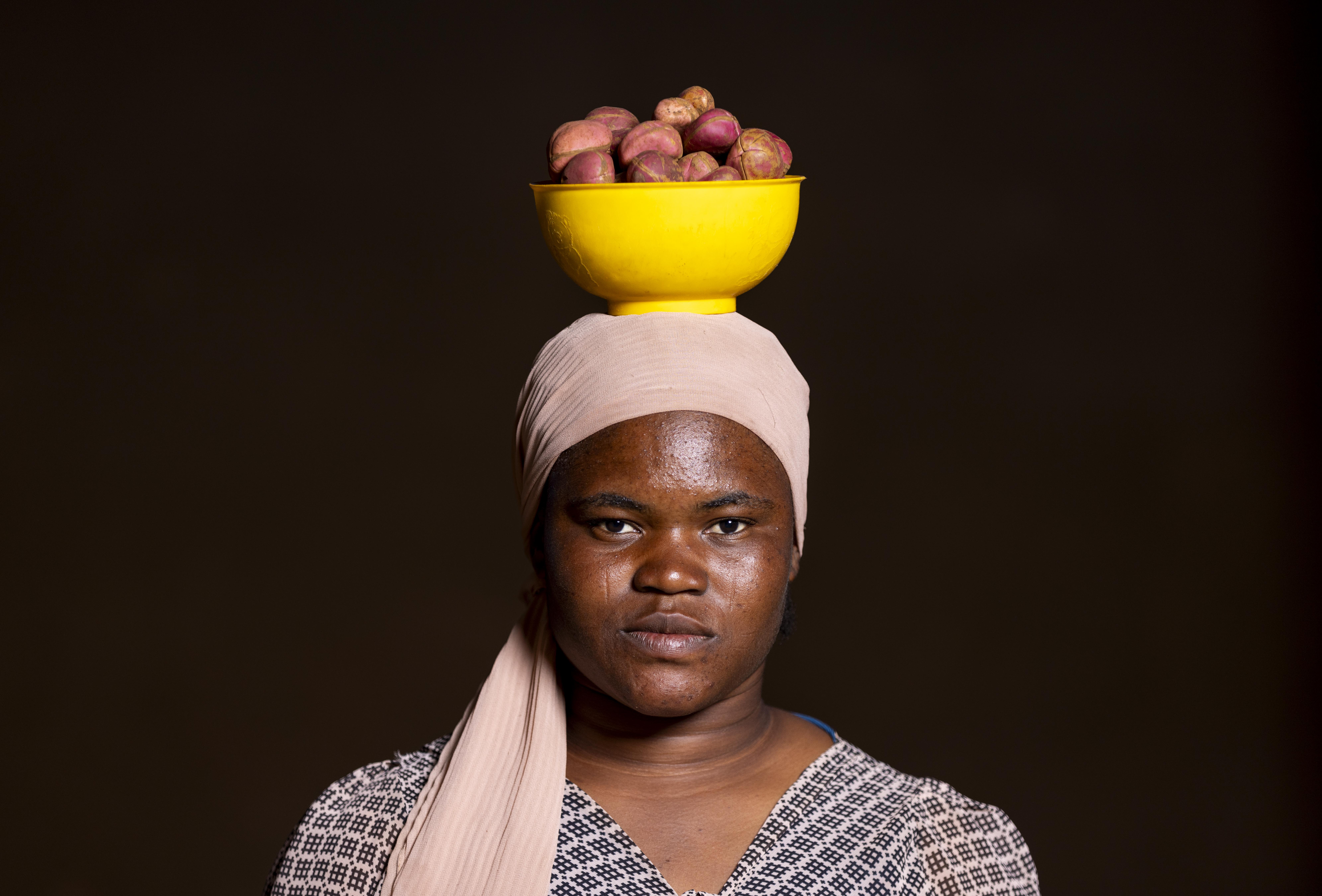 A young woman wears a patterned outfit and a headscarf and stands against a dark background. She is balancing a yellow bowl filled with kola nuts on her head. Her expression is neutral and composed.