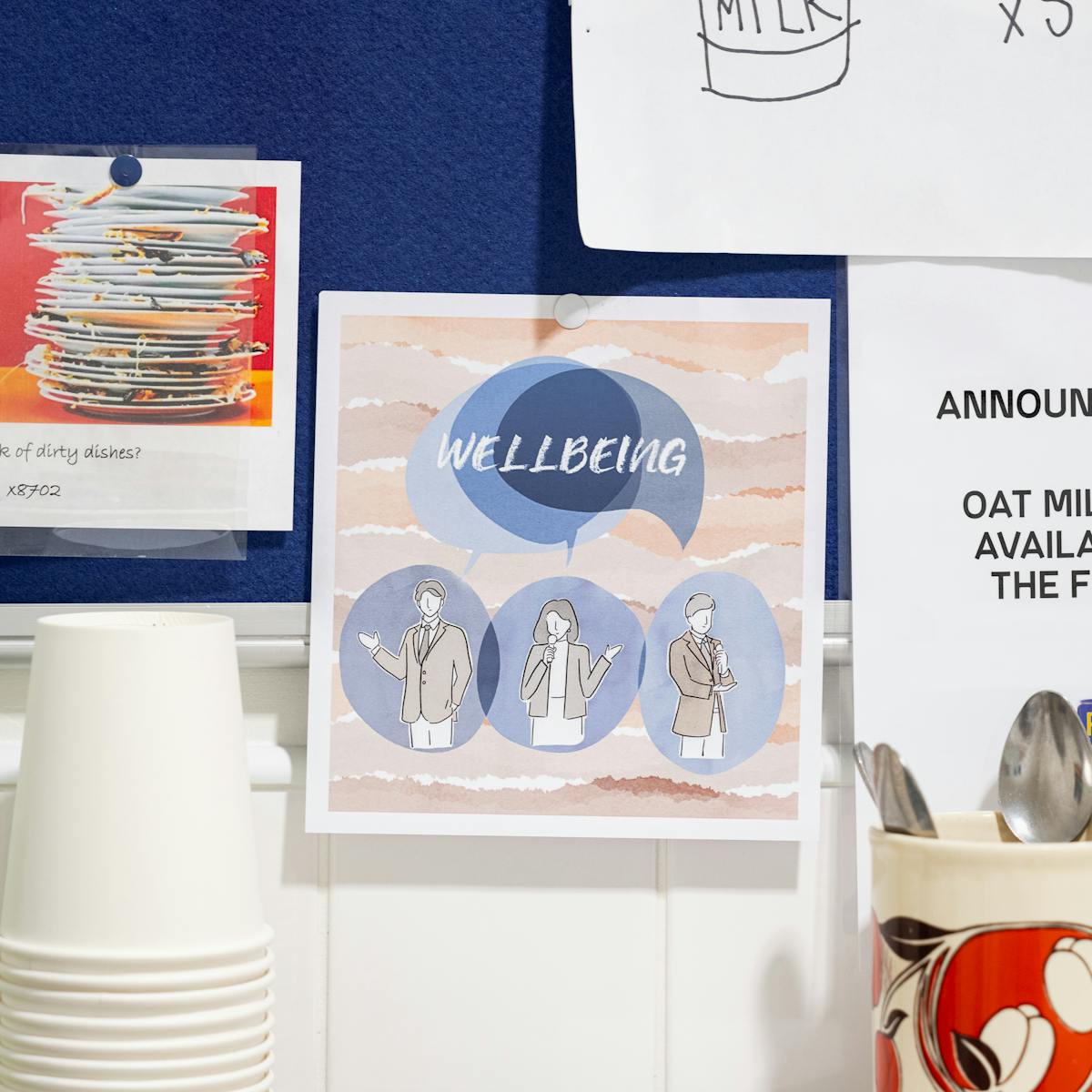 A photograph of a workplace noticeboard featuring an illustrated poster of people in an office with a speech-bubble that says Wellbeing.