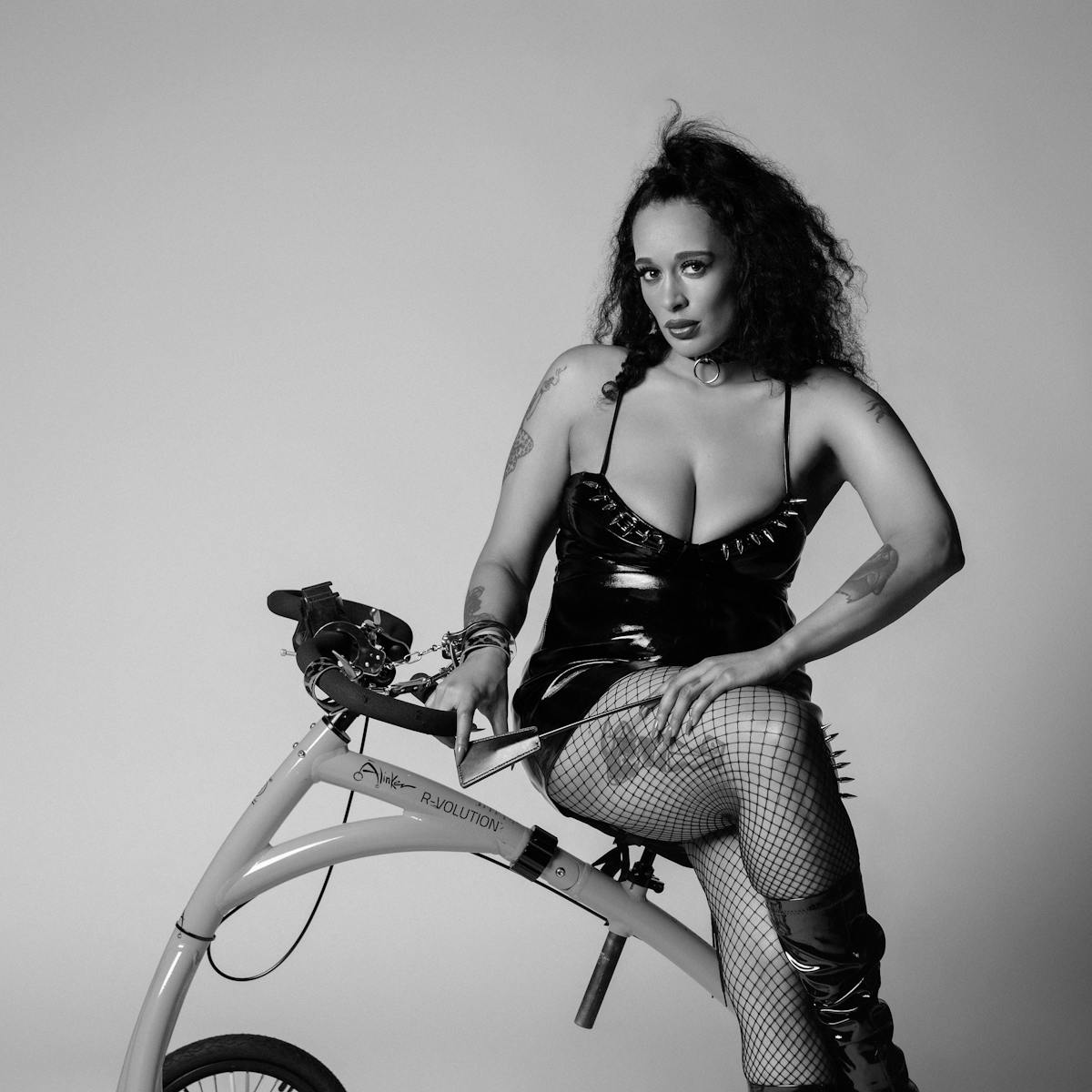 Black and white portrait of Roxy Murray dressed in a black latex mini dress with spikes and fishnet tights. She is seated side-saddle on her walking bike, her right wrist handcuffed to the handlebars while her left hand gently holds a riding crop across her lap. Roxy stares intently into the camera, lips pursed.