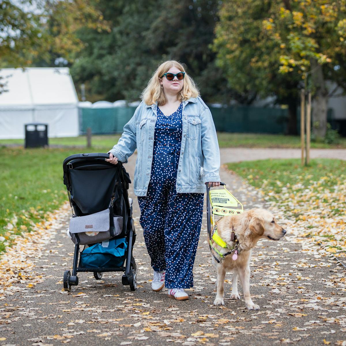 Photograph of Amy Kavanagh walking in the park, wearing a blue patterned jumpsuit, denim jacket and a pair of sunglasses. In her left hand is Amy's assistance Golden Retriever and in her right she is pulling along a children's buggy which is facing away from the camera.
