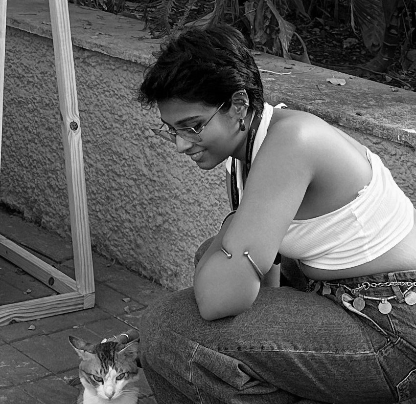 Black and white photograph of a young woman with short dark hair and glasses. She is crouching down and smiling at a cat in front of her.