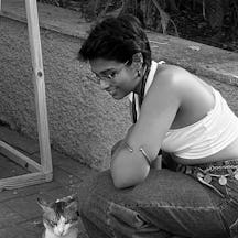 Black and white photograph of a young woman with short dark hair and glasses. She is crouching down and smiling at a cat in front of her.