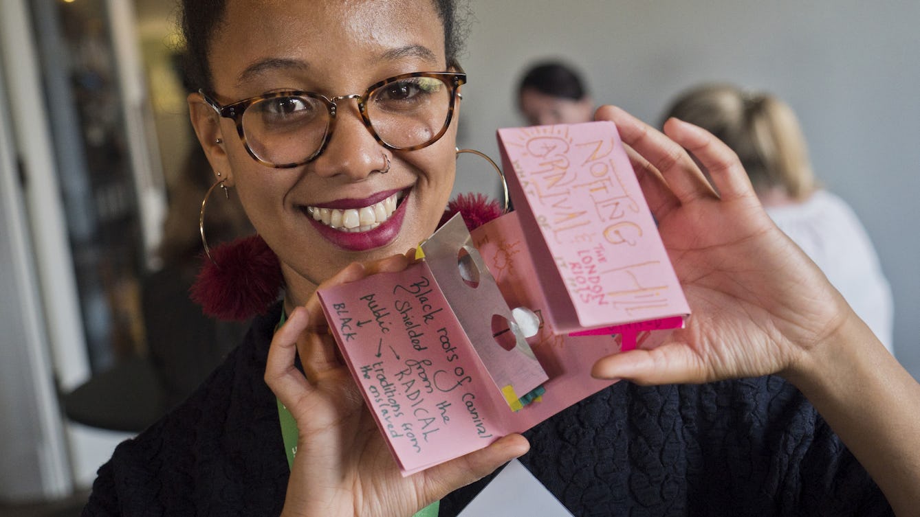 Photograph of a young woman with large round glasses and dark hair, tied back. She is smiling and looking straight at the camera while holding up a small folded paper document of a zine that she has made.