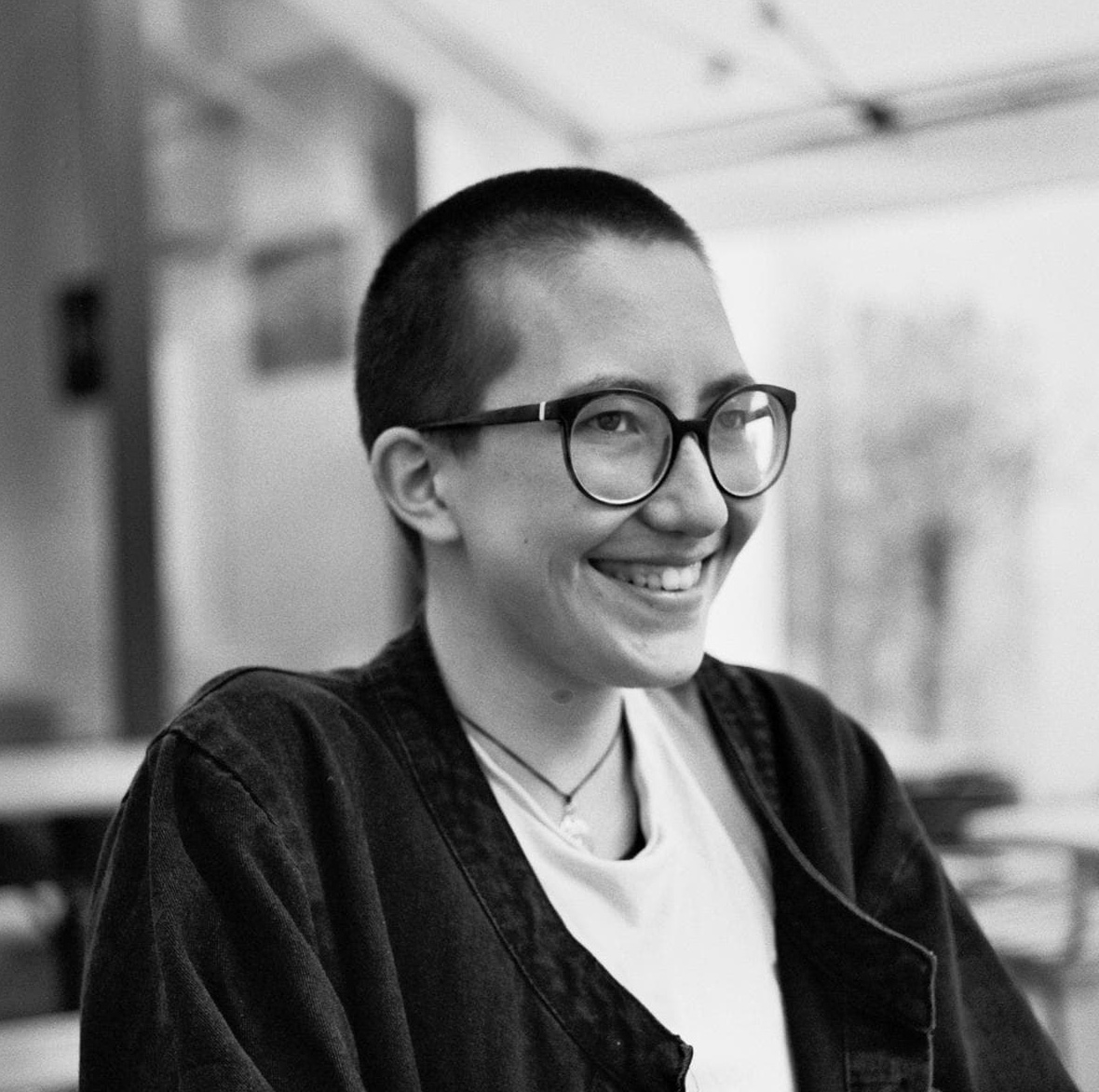 Black and white photograph of the top half of a young person with short cropped hair and glasses. They are smiling and looking at the viewer, side on.
