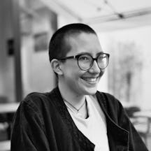 Black and white photograph of the top half of a young person with short cropped hair and glasses. They are smiling and looking at the viewer, side on.