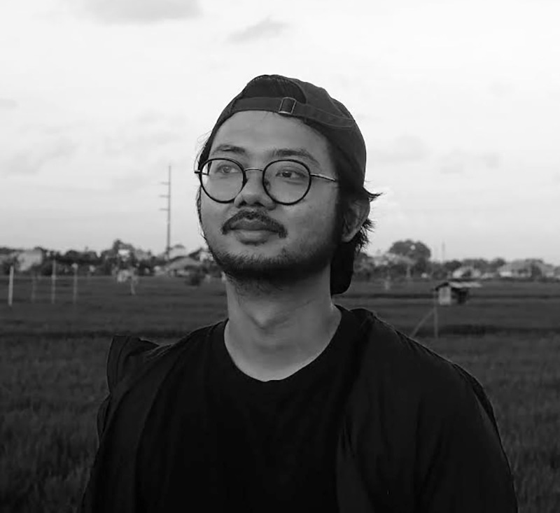 Black and white photograph of the head and shoulders of a young man standing in a field. He is wearing glasses and has a light beard and moustache.