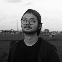 Black and white photograph of the head and shoulders of a young man standing in a field. He is wearing glasses and has a light beard and moustache.