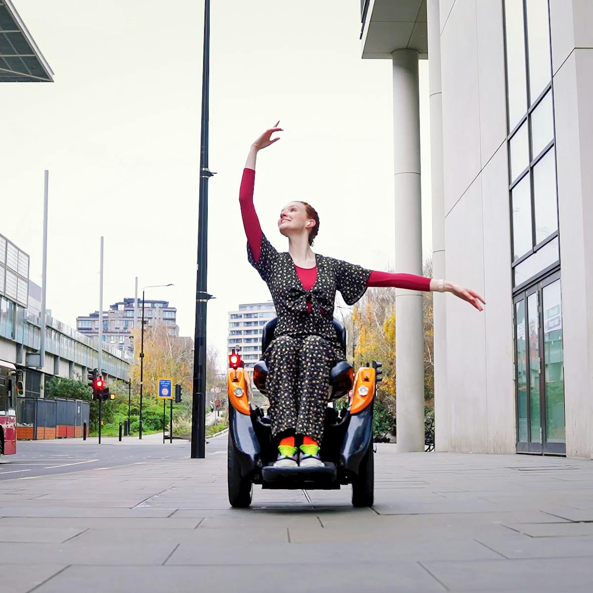 Photograph of a young woman seated in a self-balancing two-wheeled mobility device with orange accent colours, on the pavement of a busy street in Camden, London. She is wearing a flora jump suit over a long-sleeved burgundy top. Her arms are raised in a balletic pose, one arching above her, the other out to the side. Her neck is extended, chin raise, broad smile on her face.