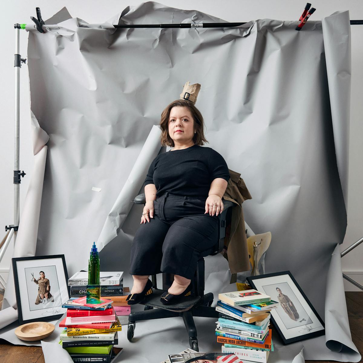 Photograph of Sinéad Burke sat on chair in the middle of a photographic studio setup where the background stands can be seen, supporting a crumpled, folded and torn grey paper background. Surrounding Sinéad at her feet at stacks of books, framed photographs, souvenirs and awards. Dressed all in black, Sinéad looks directly to camera wearing a crown cut from the cuff of a Burberry trenchcoat.