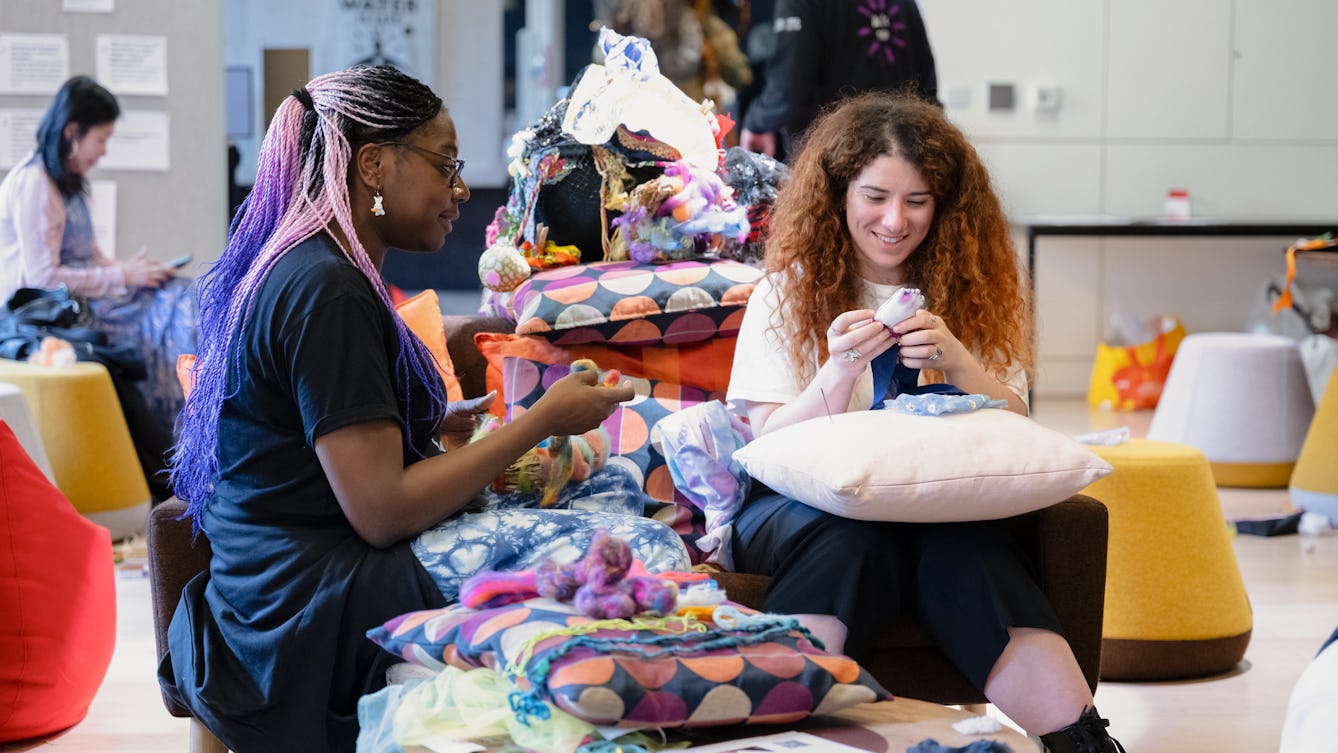 Two people at an event, sitting and engaged in crafts with textiles.