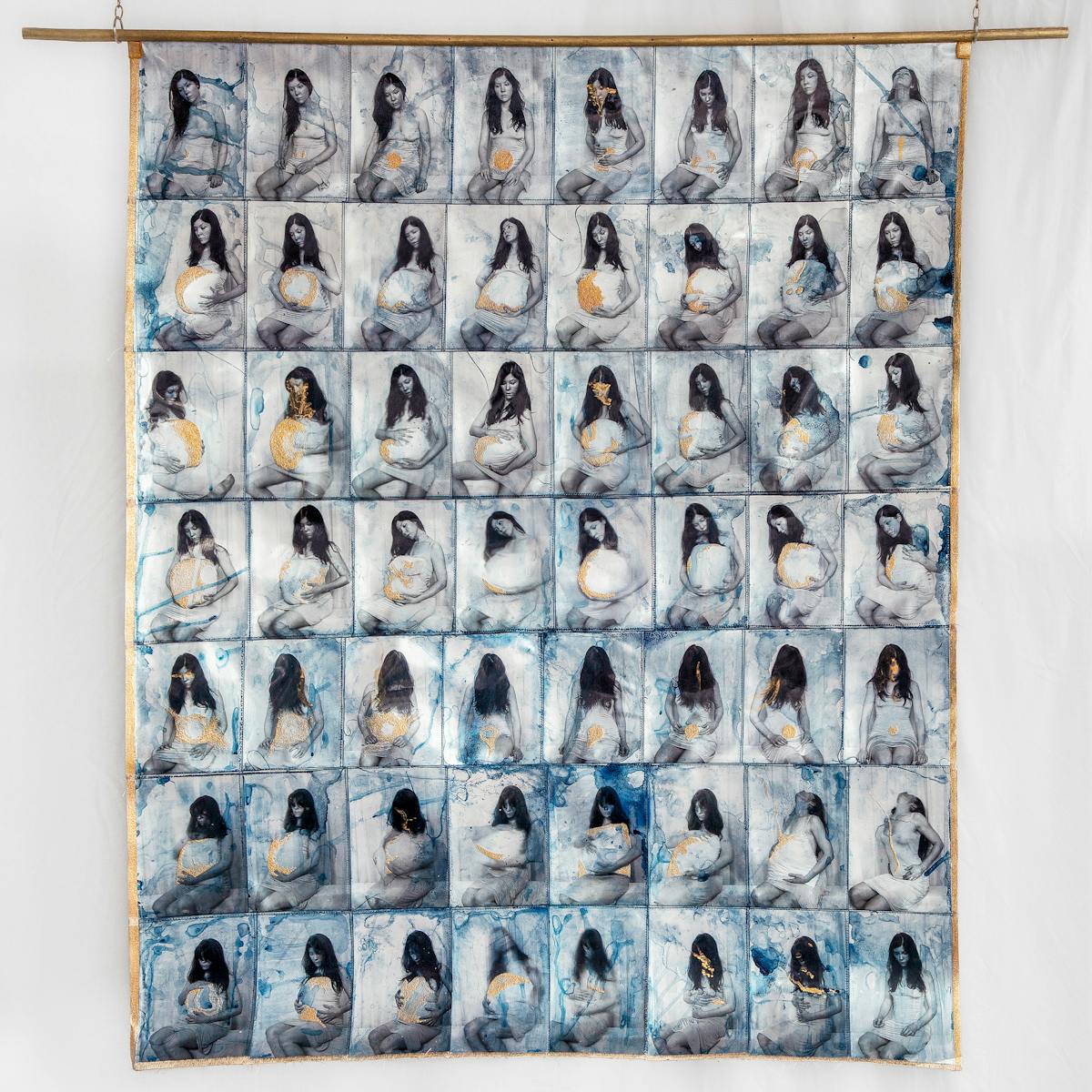 Photograph of a fabric artwork hanging from a golden pole suspended from chains. The fabric contains a grid of small photographs 8 across by 7 down. Each image shows a woman wearing a white slip with a pillow pushed up underneath, portraying her in various stages of pregnancy. Each image is embellished with gold textured paint and threaded stitches.